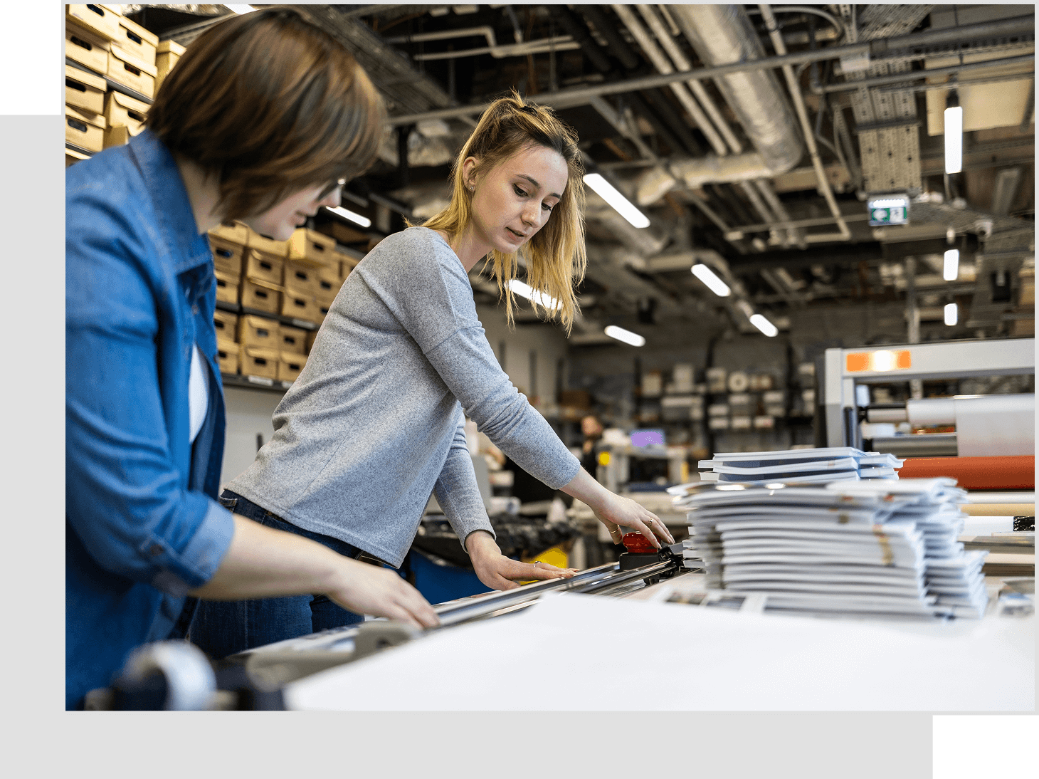 Two women working in an industrial setting, examining documents and machinery, surrounded by papers and storage boxes.