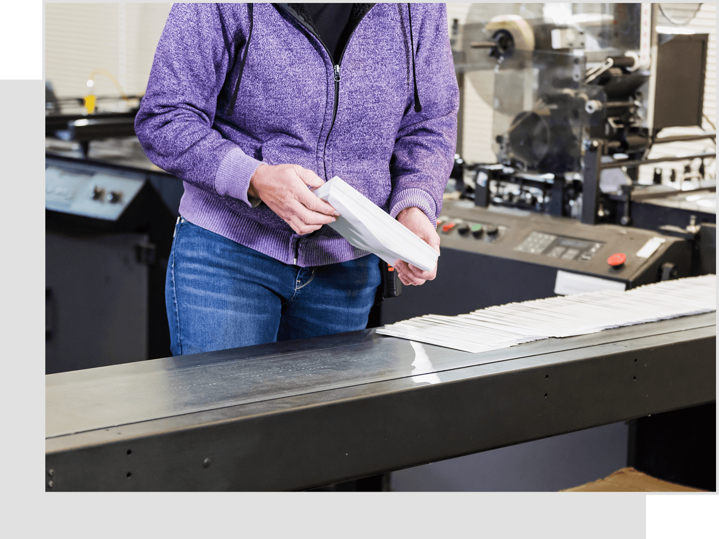 Person in a purple hoodie handling stacks of envelopes beside a conveyor belt in a factory setting.